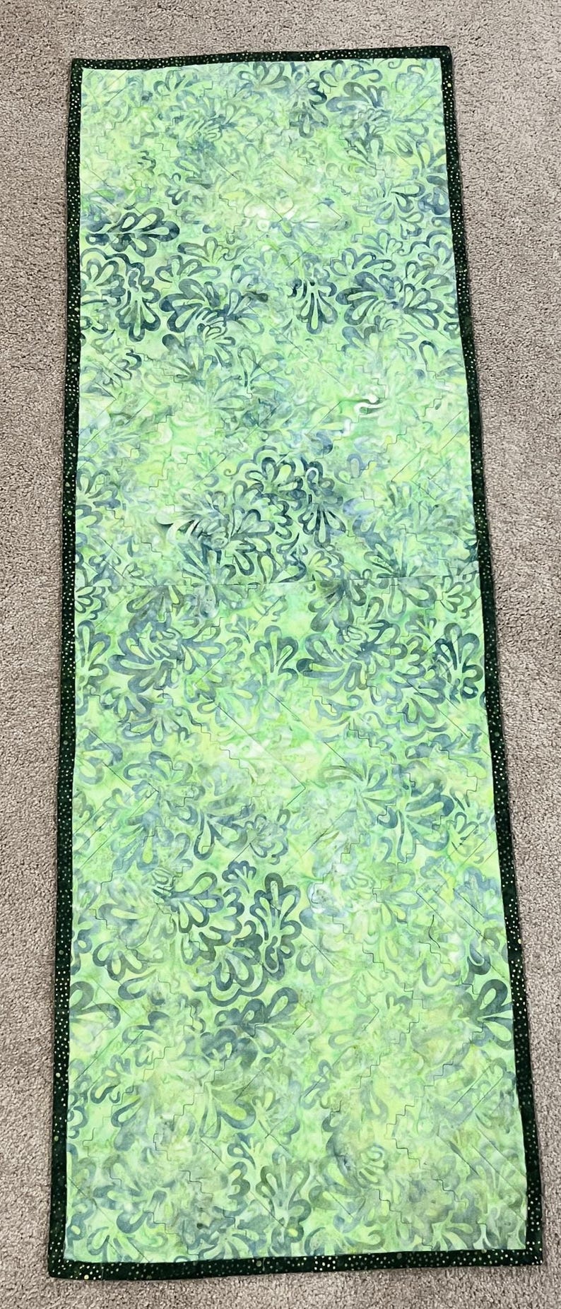 Pu&ograve; includere: Un runner da tavola rettangolare con un motivo floreale in stile batik nei toni del verde chiaro e scuro. Il tessuto &egrave; trapuntato con linee di cucitura diagonali e rifinito con un bordo verde scuro caratterizzato da piccole macchie chiare.