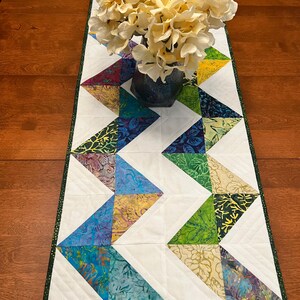 Pu&ograve; includere: Un runner da tavola trapuntato rettangolare caratterizzato da un motivo a zigzag di triangoli in tessuto batik multicolore intervallati da triangoli in tessuto bianco, rifinito con un bordo verde scuro, sormontato da un vaso di fiori artificiali color crema.