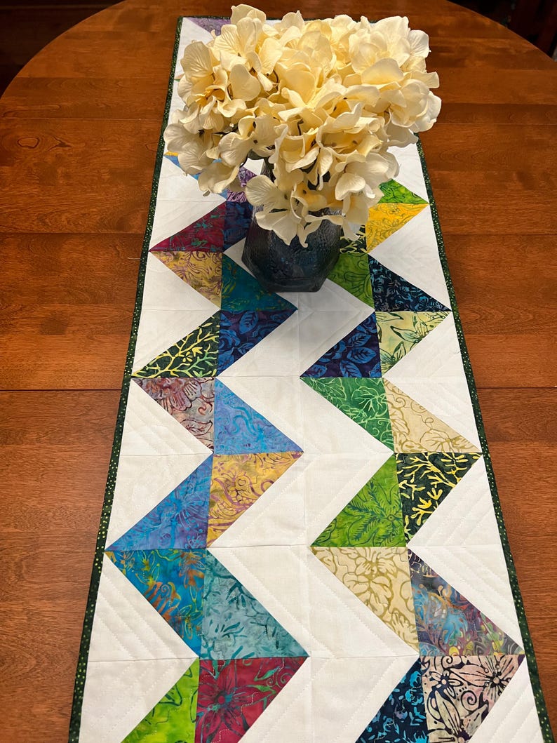 Pu&ograve; includere: Un runner da tavola trapuntato rettangolare caratterizzato da un motivo a zigzag di triangoli in tessuto batik multicolore intervallati da triangoli in tessuto bianco, rifinito con un bordo verde scuro, sormontato da un vaso di fiori artificiali color crema.