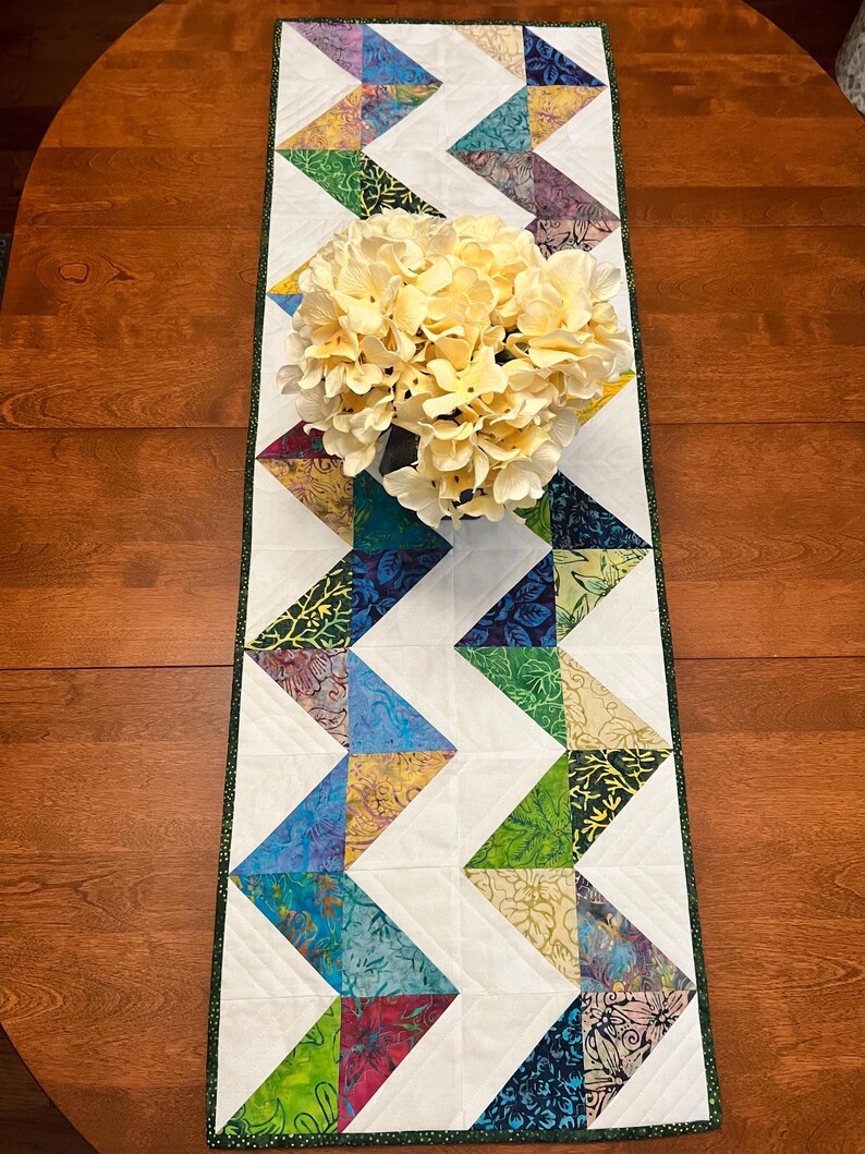 Pu&ograve; includere: Un runner da tavola trapuntato rettangolare caratterizzato da un motivo a zigzag di triangoli in tessuto batik multicolore intervallati da triangoli bianchi a tinta unita. Il runner ha un bordo verde scuro e un bouquet di fiori color crema.