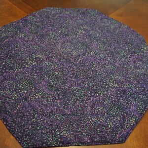 May include: An octagonal fabric mat featuring a dark purple base color with a dense, repeating pattern of swirling shapes in shades of lighter purple, teal, and black. The textile surface has a textured appearance with visible stitching along the edges.