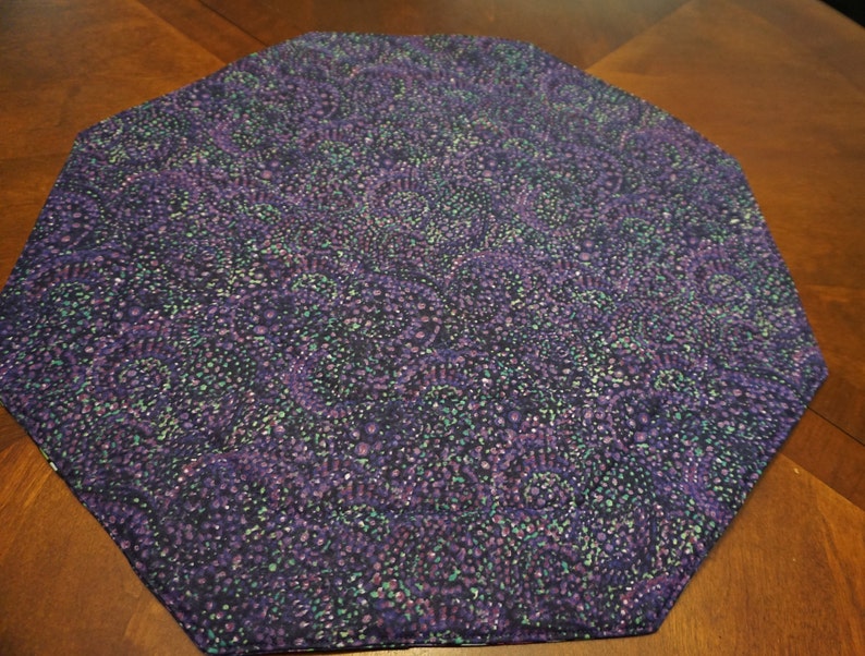 May include: An octagonal fabric mat featuring a dark purple base color with a dense, repeating pattern of swirling shapes in shades of lighter purple, teal, and black. The textile surface has a textured appearance with visible stitching along the edges.