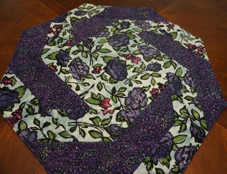 May include: A hexagonal fabric table topper featuring a spiral quilt pattern. The design alternates between a light blue fabric printed with dark purple roses and green leaves, and a dark purple fabric with a dense, speckled texture pattern.