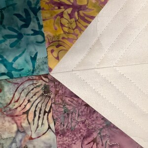 Pu&ograve; includere: Un pezzo di tessuto trapuntato con sezioni triangolari in motivi batik turchese, giallo e viola accanto a una sezione triangolare bianca unita con cuciture diagonali. Un bordo verde scuro con piccoli punti gialli rifinisce il lato.