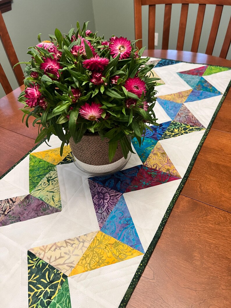 Pu&ograve; includere: Un runner da tavola rettangolare caratterizzato da un design patchwork geometrico con triangoli di tessuto bianco che si alternano a triangoli di tessuto batik multicolore nelle tonalit&agrave; del blu, verde, giallo e viola, rifinito con un bordo verde scuro maculato.