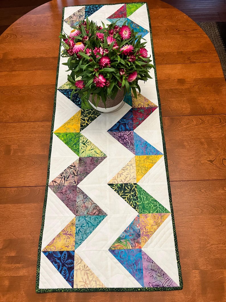 Pu&ograve; includere: Un runner da tavola trapuntato rettangolare caratterizzato da un vivace motivo a zigzag realizzato con triangoli di tessuto batik multicolore nelle tonalit&agrave; del blu, viola, giallo e verde, a contrasto con sezioni bianche e un bordo verde scuro.
