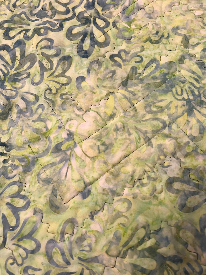 Pu&ograve; includere: Un tessuto trapuntato con un motivo in stile batik caratterizzato da forme floreali astratte in tonalit&agrave; verde chiaro, lime e blu tenue. Il tessuto &egrave; cucito con un distinto motivo a zigzag che crea una griglia geometrica sulla superficie.
