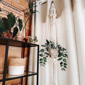 Puede incluir: Un macetero de macramé blanco con una cesta tejida que contiene una planta verde. El macetero está colgado de una barra de cortina delante de una cortina blanca. El macetero está en una sala de estar con una estantería y una pared de ladrillo.