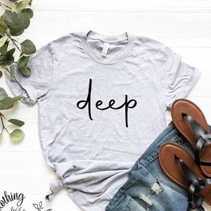 May include: A heather grey t-shirt with the word "deep" written in black cursive script. The shirt is laid out on a white wooden surface with a pair of brown sandals and blue jeans.