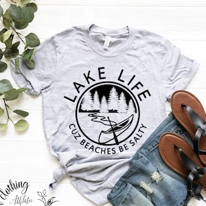 Puede incluir: Una camiseta gris con un gr&aacute;fico circular negro que dice "Lake Life Cuz Beaches Be Salty". El gr&aacute;fico presenta una canoa en un lago con &aacute;rboles en el fondo.