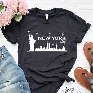 May include: Black t-shirt with a white graphic of the New York City skyline, including the Statue of Liberty and the Twin Towers. The text "NEW YORK city" is printed on the shirt.