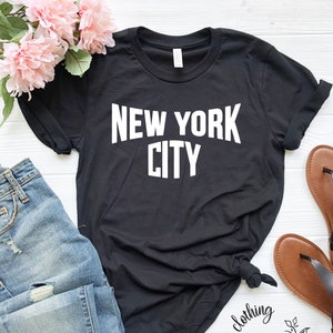 May include: Black t-shirt with white text that reads "NEW YORK CITY". The shirt is shown on a white surface with blue jeans and brown sandals.