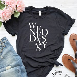 May include: Black t-shirt with white text that reads "WE NED DAS Y". The shirt is shown on a white surface with a pair of brown sandals and blue jeans.