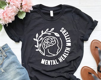 Puede incluir: Camiseta negra con un gráfico blanco de un cerebro dentro de un círculo con hojas y el texto "Mental Health Matters".