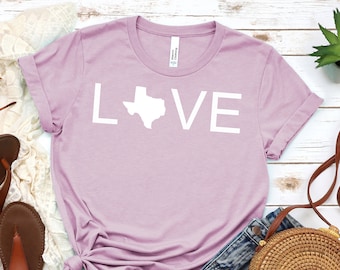 May include: A lavender t-shirt with the word "LOVE" printed in white, with the letter "O" replaced by the outline of the state of Texas. The shirt is laid out on a white wooden surface with a pair of brown sandals, a pair of denim shorts, a brown woven bag, and a small potted succulent plant.
