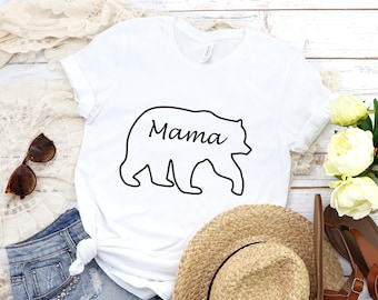 May include: White t-shirt with a black outline of a bear and the word "Mama" inside the bear. The shirt is laying on a white wooden surface with a pair of denim shorts, a straw hat, sunglasses, and a bouquet of white flowers.
