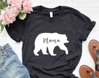 May include: Black t-shirt with a white silhouette of a bear and the word "Mama" printed on it. The shirt is laid flat on a white surface with a pair of brown sandals and a pair of blue jeans.