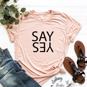 May include: A light pink t-shirt with the text "SAY YES" printed in black. The shirt is laid flat on a white surface with a pair of brown sandals and a pair of blue jeans.