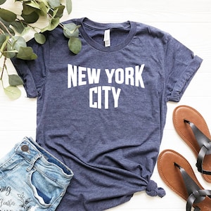 May include: A navy blue t-shirt with the text "NEW YORK CITY" in white lettering. The shirt is laid flat on a white surface with a pair of brown sandals and denim shorts.