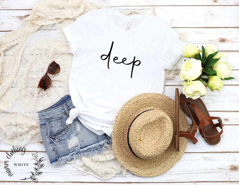 May include: White t-shirt with the word "deep" written in black cursive script. The shirt is laid out on a white wooden surface with a straw hat, denim shorts, sunglasses, and a bouquet of white peonies.
