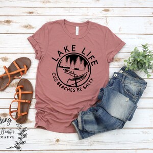 Puede incluir: Una camiseta de color malva brezo con un dise&ntilde;o gr&aacute;fico negro que dice "Lake Life Cuz Beaches Be Salty" con una canoa y &aacute;rboles en un c&iacute;rculo.