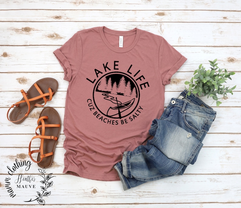 Puede incluir: Una camiseta de color malva brezo con un dise&ntilde;o gr&aacute;fico negro que dice "Lake Life Cuz Beaches Be Salty" con una canoa y &aacute;rboles en un c&iacute;rculo.