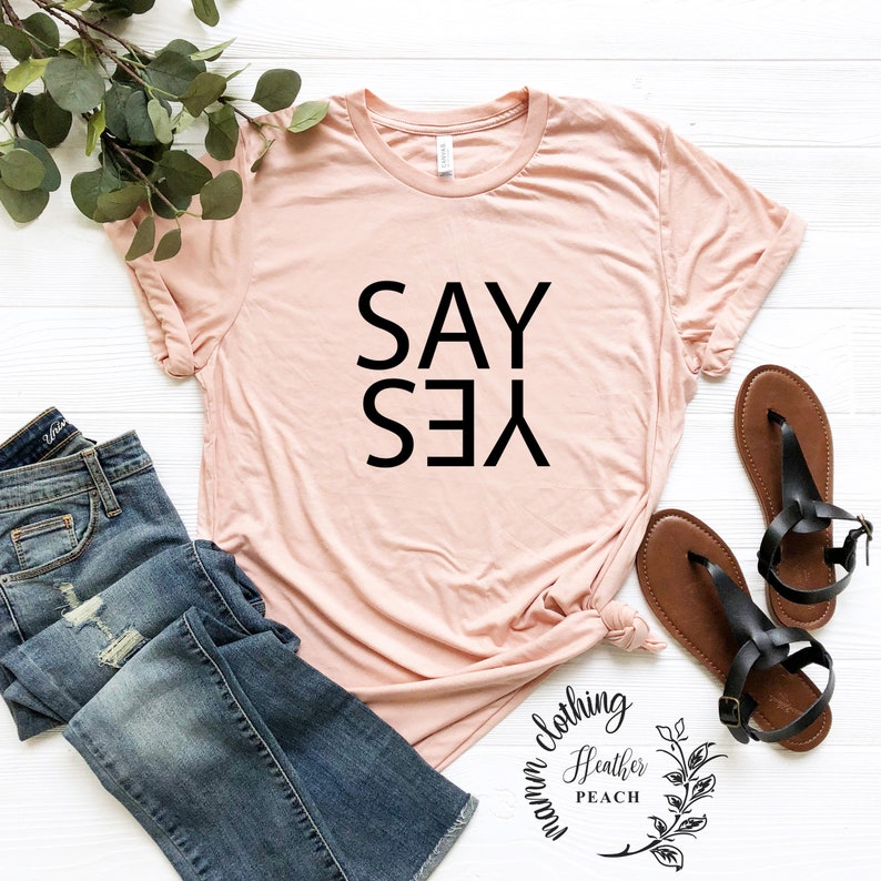 May include: A light pink t-shirt with the text "SAY YES" printed in black. The shirt is laid flat on a white surface with a pair of brown sandals and a pair of blue jeans.