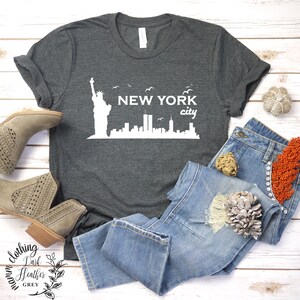 May include: A dark heather grey t-shirt with a white graphic of the Statue of Liberty and the New York City skyline. The text "NEW YORK city" is printed below the graphic.