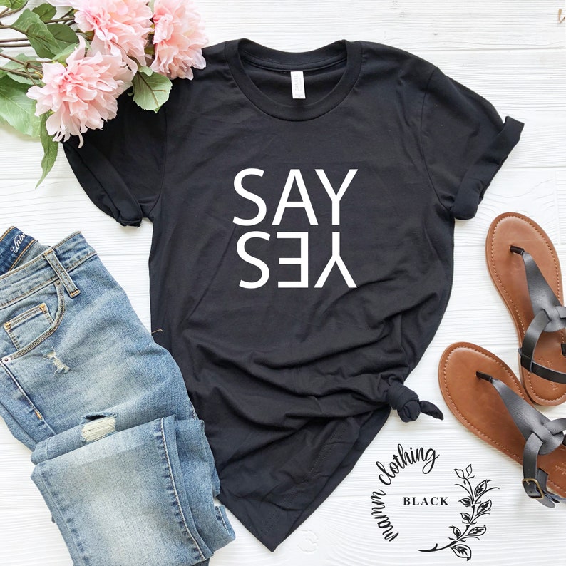 May include: Black t-shirt with white text that reads "SAY YES" in a vertical orientation. The shirt is laid flat on a white surface with a pair of brown sandals and blue jeans.