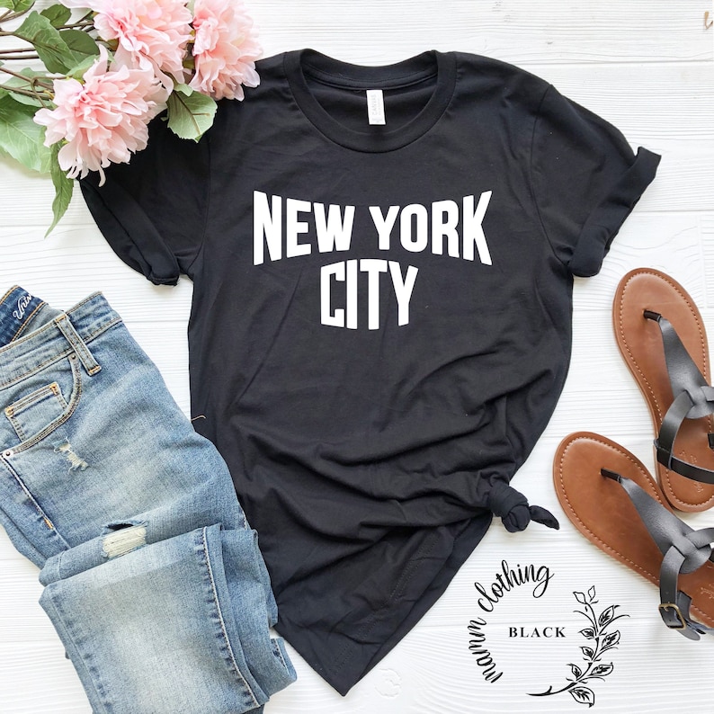 May include: Black t-shirt with white text that reads "NEW YORK CITY". The shirt is shown on a white surface with blue jeans and brown sandals.