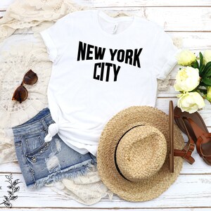 May include: White t-shirt with black text that reads "NEW YORK CITY". The shirt is laid flat on a white wooden surface with a straw hat, denim shorts, sunglasses, and a bouquet of white flowers.