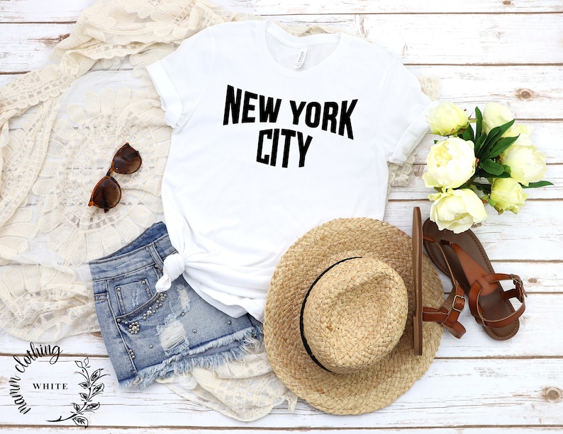 May include: White t-shirt with black text that reads "NEW YORK CITY". The shirt is laid flat on a white wooden surface with a straw hat, denim shorts, sunglasses, and a bouquet of white flowers.
