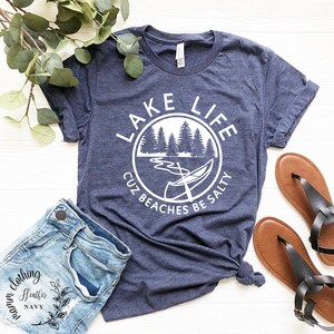 Puede incluir: Una camiseta azul marino con un gr&aacute;fico circular blanco que dice "Lake Life Cuz Beaches Be Salty". El gr&aacute;fico presenta una canoa en un lago con &aacute;rboles en el fondo.
