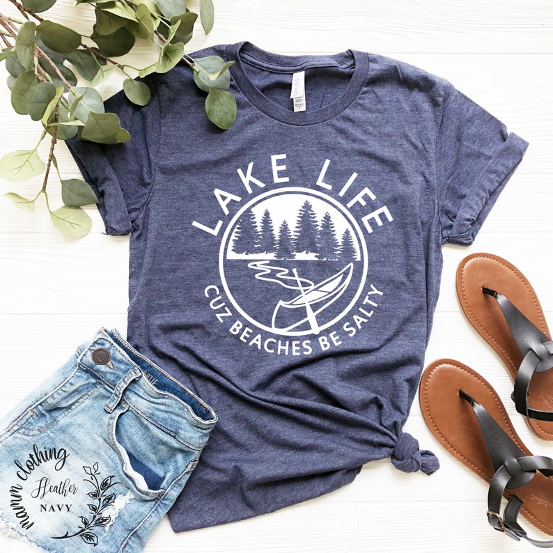 Puede incluir: Una camiseta azul marino con un gr&aacute;fico circular blanco que dice "Lake Life Cuz Beaches Be Salty". El gr&aacute;fico presenta una canoa en un lago con &aacute;rboles en el fondo.