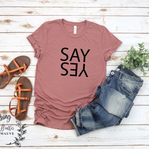 May include: A heather mauve t-shirt with the words "SAY YES" printed in black.