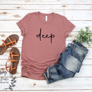 May include: A heather mauve t-shirt with the word "deep" written in black cursive script. The shirt is photographed on a white wood background with a pair of brown sandals, blue jeans, and a small potted plant.