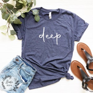May include: A navy blue t-shirt with the word "deep" written in white cursive script. The shirt is shown on a white wooden surface with a pair of brown sandals and a pair of blue denim shorts.