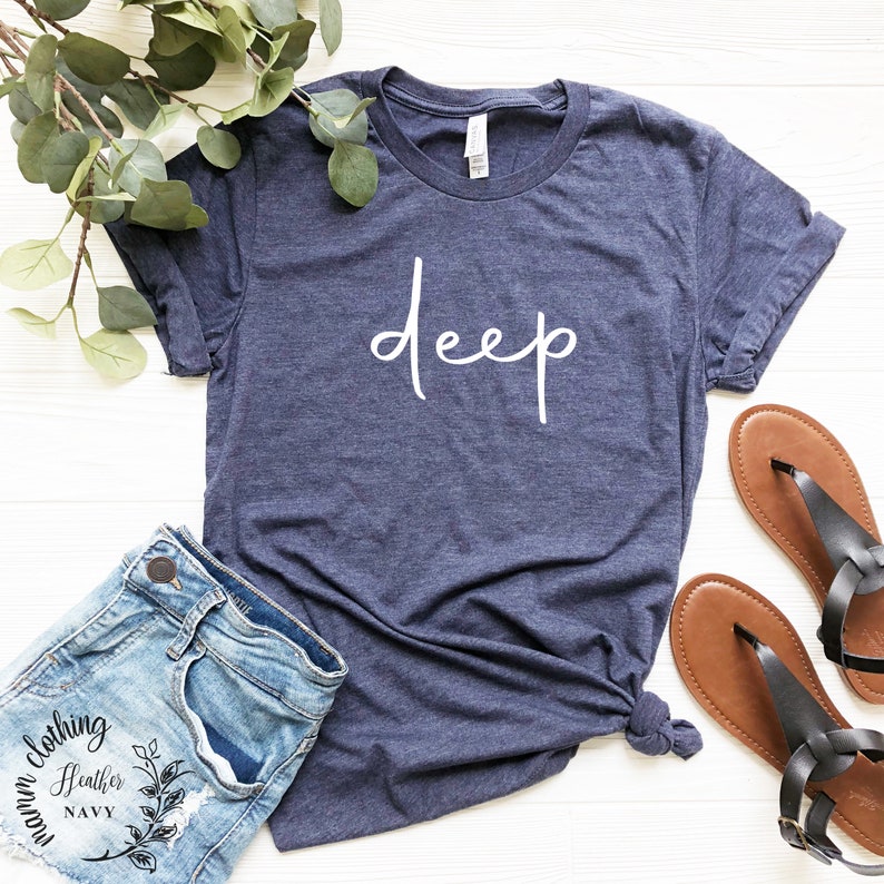 May include: A navy blue t-shirt with the word "deep" written in white cursive script. The shirt is shown on a white wooden surface with a pair of brown sandals and a pair of blue denim shorts.