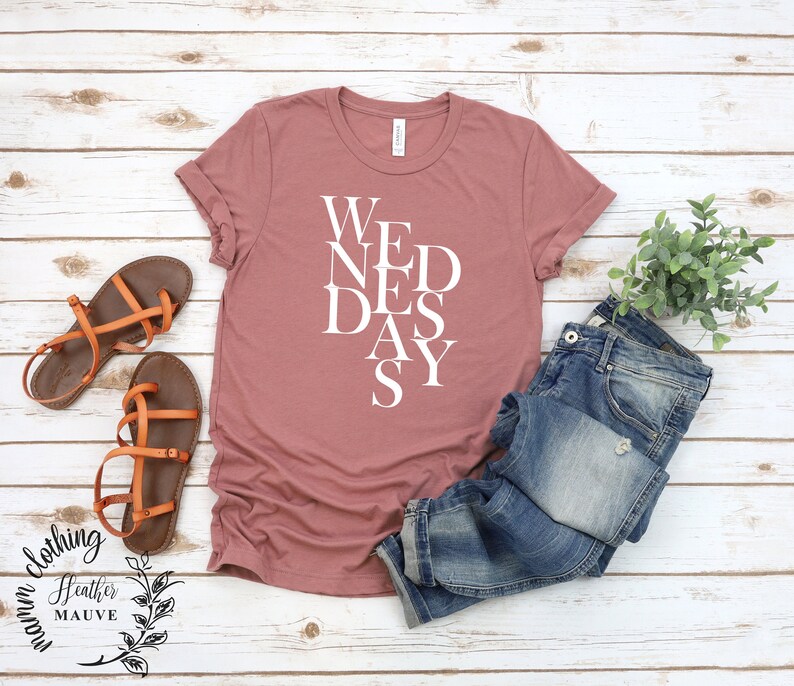 May include: A heather mauve t-shirt with the word "WEDNESDAY" printed on it in white letters, arranged in a vertical, staggered pattern. The shirt is photographed on a white wooden background with a pair of brown sandals, a pair of blue jeans, and a potted plant.