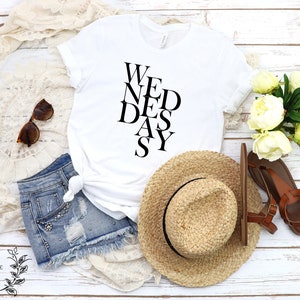 May include: White t-shirt with black text that reads "We Need a Day". The shirt is styled with denim shorts, a straw hat, and sandals.