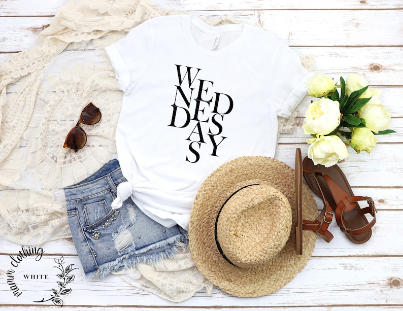 May include: White t-shirt with black text that reads "We Need a Day". The shirt is styled with denim shorts, a straw hat, and sandals.