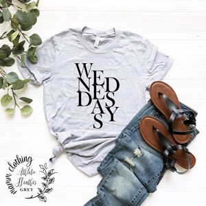 May include: A gray t-shirt with the word "WEDNESDAY" spelled out in a vertical, staggered design. The shirt is on a white wooden surface with a pair of brown sandals and blue jeans.
