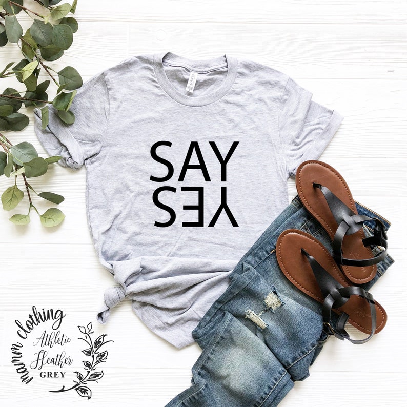 May include: A gray t-shirt with the text "SAY YES" printed in black on the front. The shirt is laid flat on a white surface with a pair of brown sandals and blue jeans.