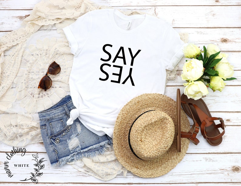 May include: White t-shirt with the text "SAY YES" printed in black on the front. The shirt is laid flat on a white wooden surface with a pair of denim shorts, a straw hat, sunglasses, and a bouquet of white flowers.