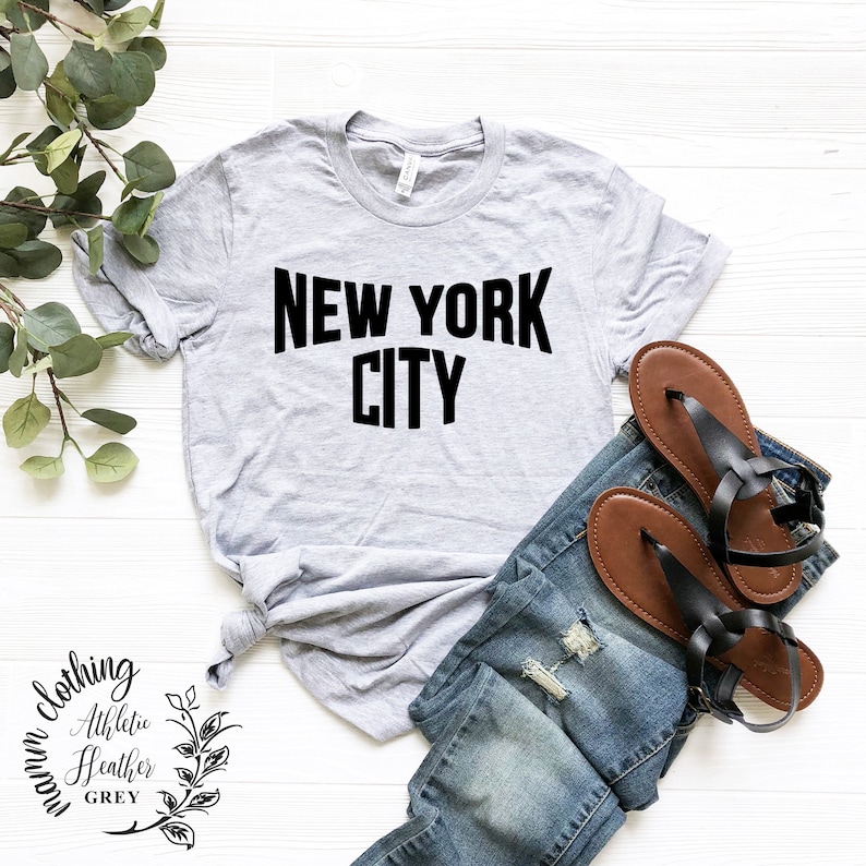 May include: A gray t-shirt with the text "NEW YORK CITY" printed in black. The shirt is laid out on a white wooden surface with a pair of brown sandals and blue jeans.