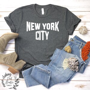 May include: A dark heather grey t-shirt with the text "NEW YORK CITY" printed in white on the front. The shirt is laid out on a white wooden surface with a pair of brown ankle boots and blue jeans.