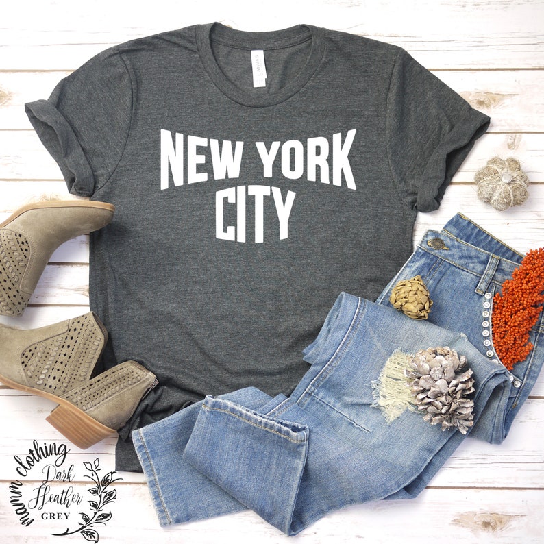 May include: A dark heather grey t-shirt with the text "NEW YORK CITY" printed in white on the front. The shirt is laid out on a white wooden surface with a pair of brown ankle boots and blue jeans.