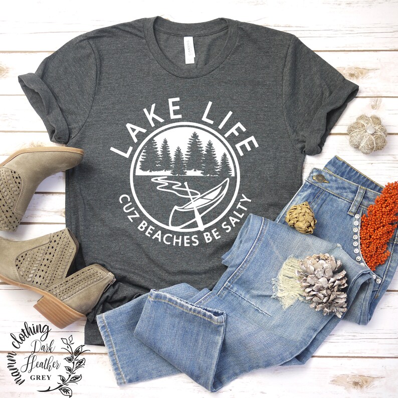 Puede incluir: Una camiseta gris oscuro jaspeada con un dise&ntilde;o gr&aacute;fico blanco que dice "Lake Life Cuz Beaches Be Salty". El gr&aacute;fico presenta una canoa, &aacute;rboles y un lago.