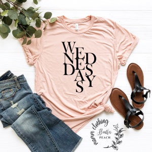 May include: A pink t-shirt with the word "WEDNESDAY" spelled out in black letters, with the letters arranged in a vertical, staggered pattern. The shirt is on a white surface with a pair of brown sandals and a pair of blue jeans.