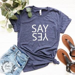 May include: A navy blue t-shirt with the words "SAY YES" printed in white on the front. The shirt is laid flat on a white surface with a pair of brown sandals and a pair of blue denim shorts.
